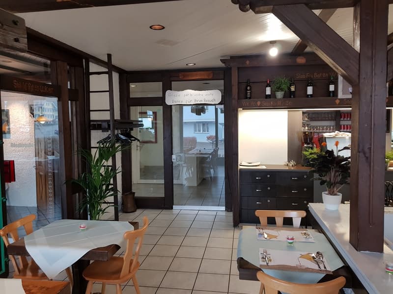 Indoor dining area with tables, chairs, wooden beams, shelves with bottles, plants, tiled floor.