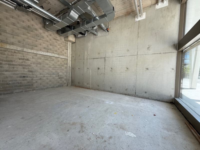 The image shows a large, open commercial space with concrete walls and floors. The space has exposed ventilation ducts and pipes running along the ceiling, indicating it is likely an industrial or warehouse-type setting. The space appears to be unfinished