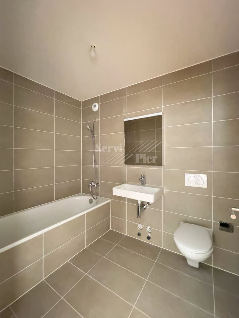 The image shows a modern bathroom with beige tile walls, a bathtub, a sink, and a toilet. The bathroom has a minimalist and clean design with a recessed mirror cabinet and a simple light fixture on the ceiling.