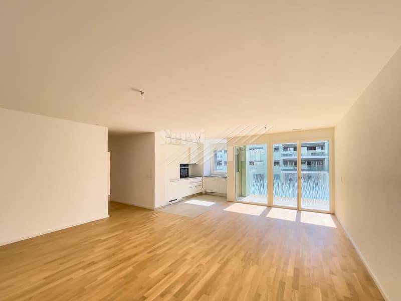 Spacious open-plan living area with hardwood flooring, large windows providing natural light, and a balcony or terrace visible. The kitchen area is integrated into the living space with modern white cabinets.