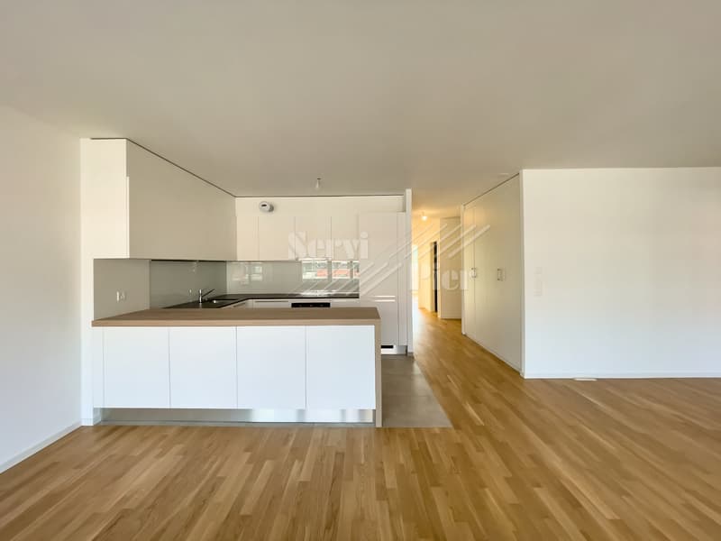 Open-concept living space with a modern kitchen featuring white cabinets, a countertop, and a dishwasher. The room has hardwood flooring and a hallway leading to other areas of the apartment.