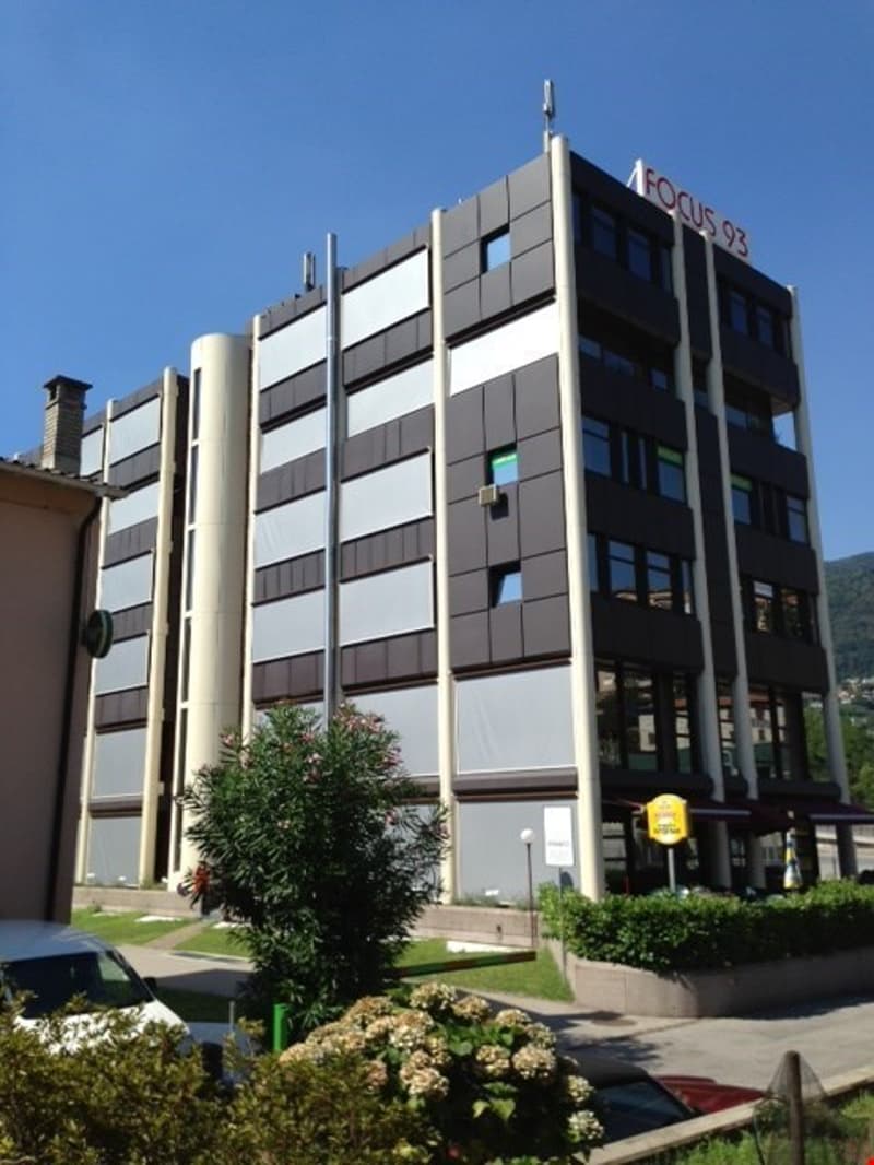 A multi-story office building with a modern, geometric facade made of glass and metal panels. The building has several floors and appears to be located in an urban setting with landscaping and parking spaces around it.