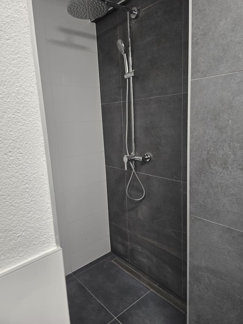 Shower area with showerhead and shower hose, grey tiled walls, tiled floor with drainage grate