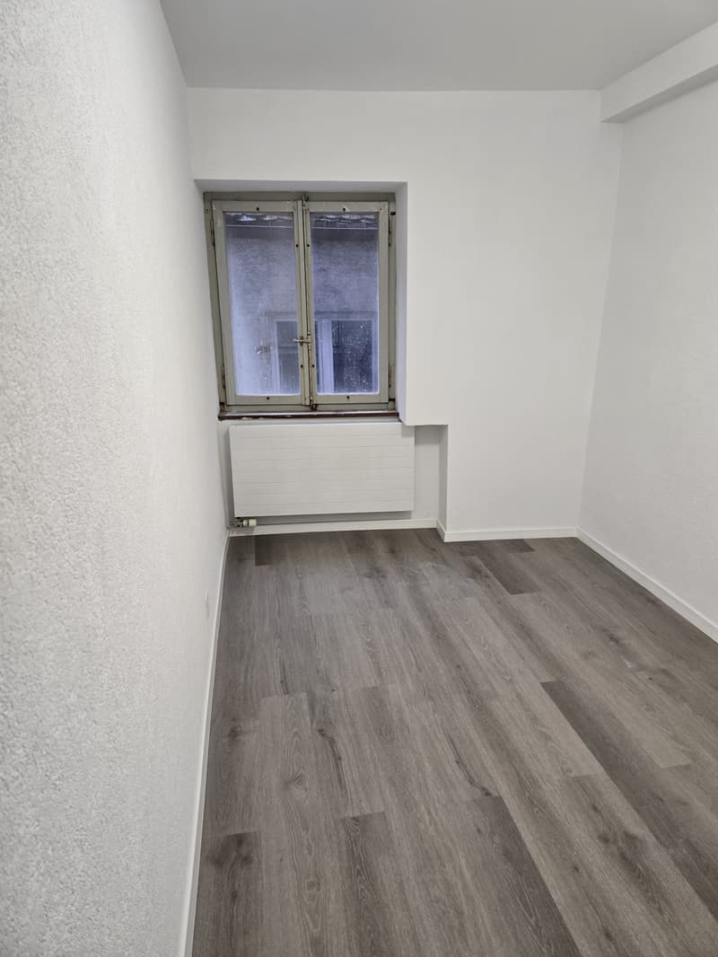 Empty room with white walls, wooden floors, and a window. There is a radiator under the window.