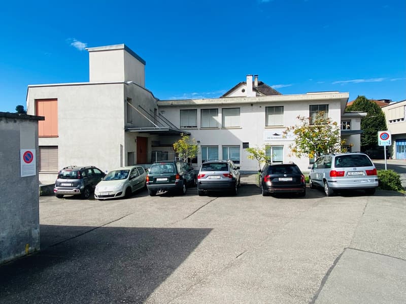 white building, several cars parked in front, concrete parking lot, trees in the background