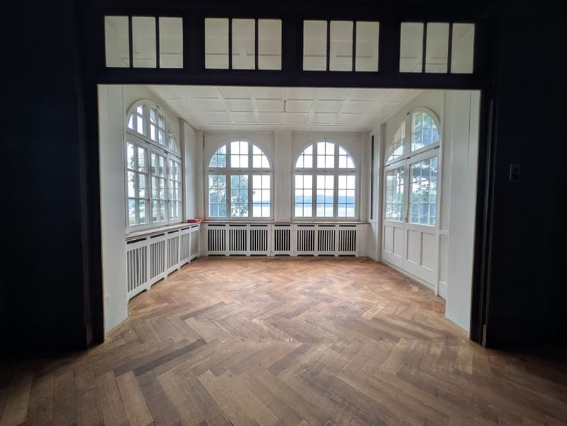 Empty room with wooden floor, white walls, large arched windows with views of the outside.