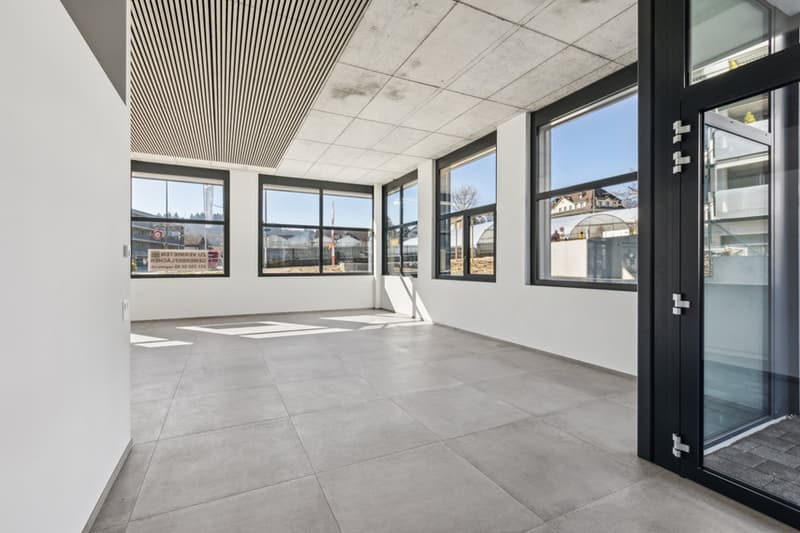 Large open commercial space with high ceilings, large windows, and a modern, minimalist design. The space features a concrete floor and a suspended ceiling with linear lighting fixtures. The windows provide ample natural light and views of the surrounding