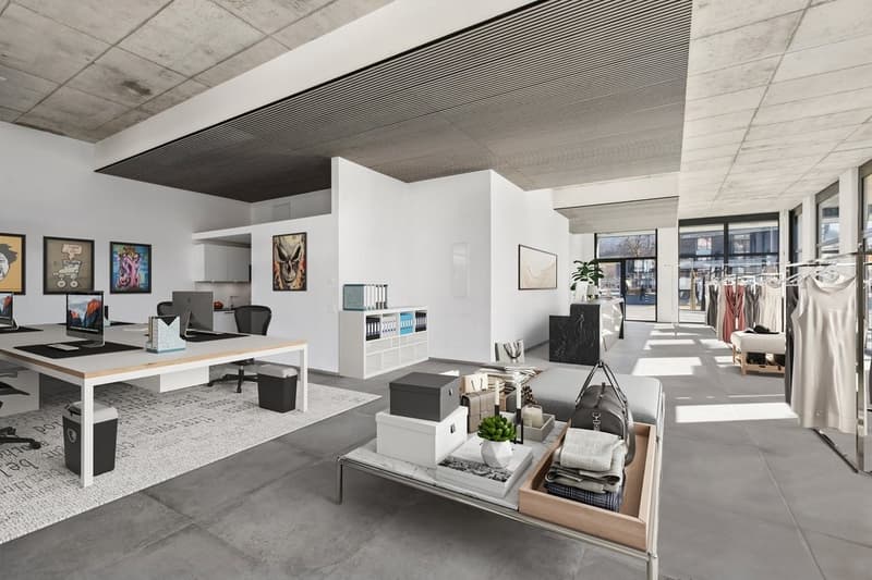The image shows a modern, open-plan office space with a minimalist design. The space features white walls, a concrete ceiling, and large windows that provide natural light. There are several workstations with desks, computers, and office equipment. The sp