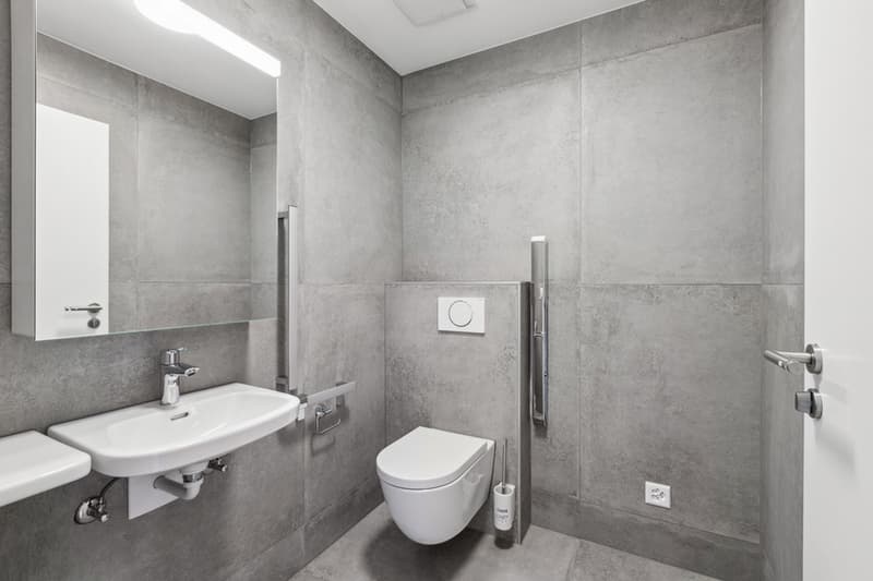Large bathroom with grey tiled walls, a white sink and toilet, and a shower stall.