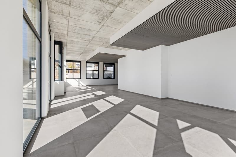 Large open-plan office space with concrete ceiling, gray tiled flooring, and large windows providing ample natural light. The space has a modern, minimalist design aesthetic.