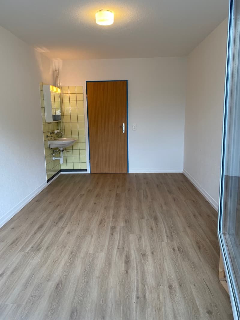 an empty room with wooden floor, bathroom tiles, sink, mirror, white walls, and a wooden door