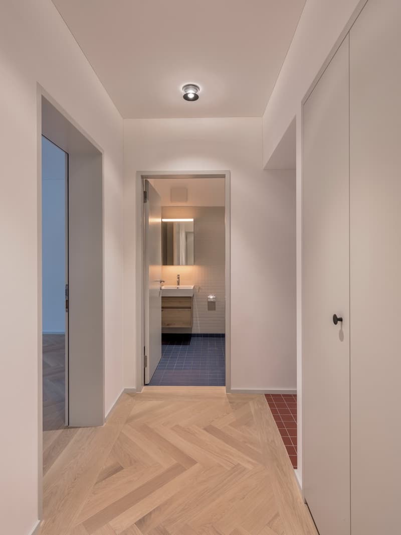Bathroom: open hallway, white walls, wooden floor, small sink, mirror, white tiled wall, blue tiled floor, toilet