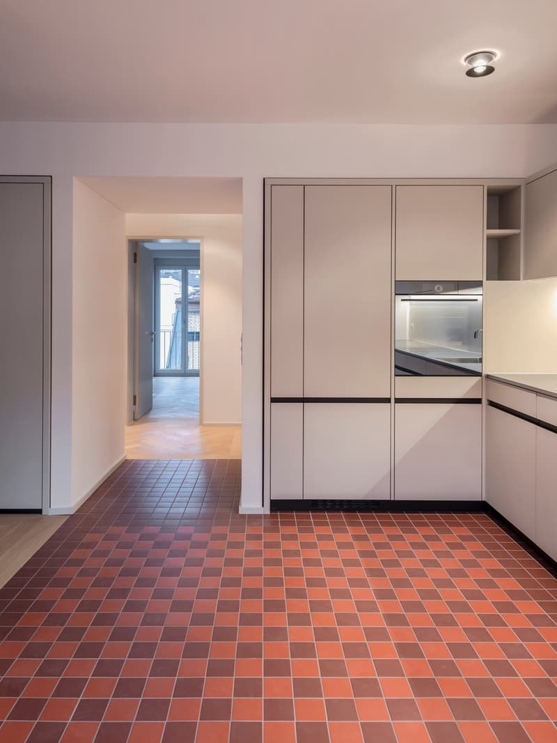modern kitchen with large window, wooden floor, orange and brown tiled floor, white cabinets, integrated oven and microwave