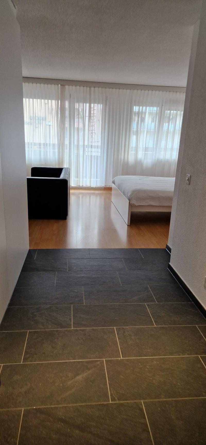 Empty room with wooden floor, white walls, large windows, black sofa, white bed