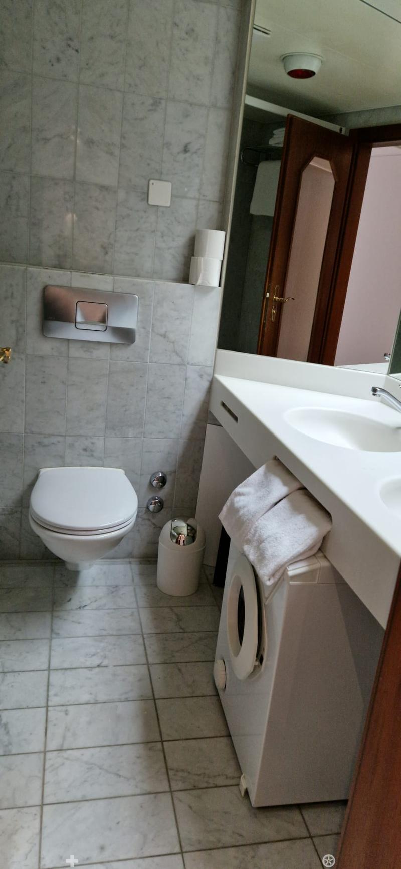 a small bathroom with white tiled floor, white ceramic toilet, white ceramic sink, a mirror, a washing machine with a towel placed on it