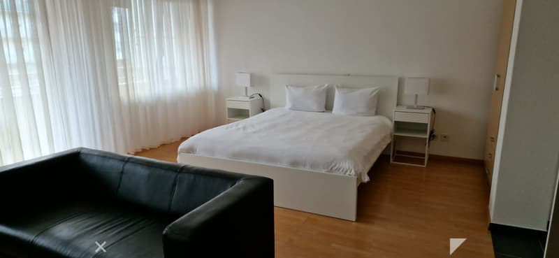 A bedroom with white bed, two white nightstands with lamps, white pillows, black leather couch, and wooden floor.