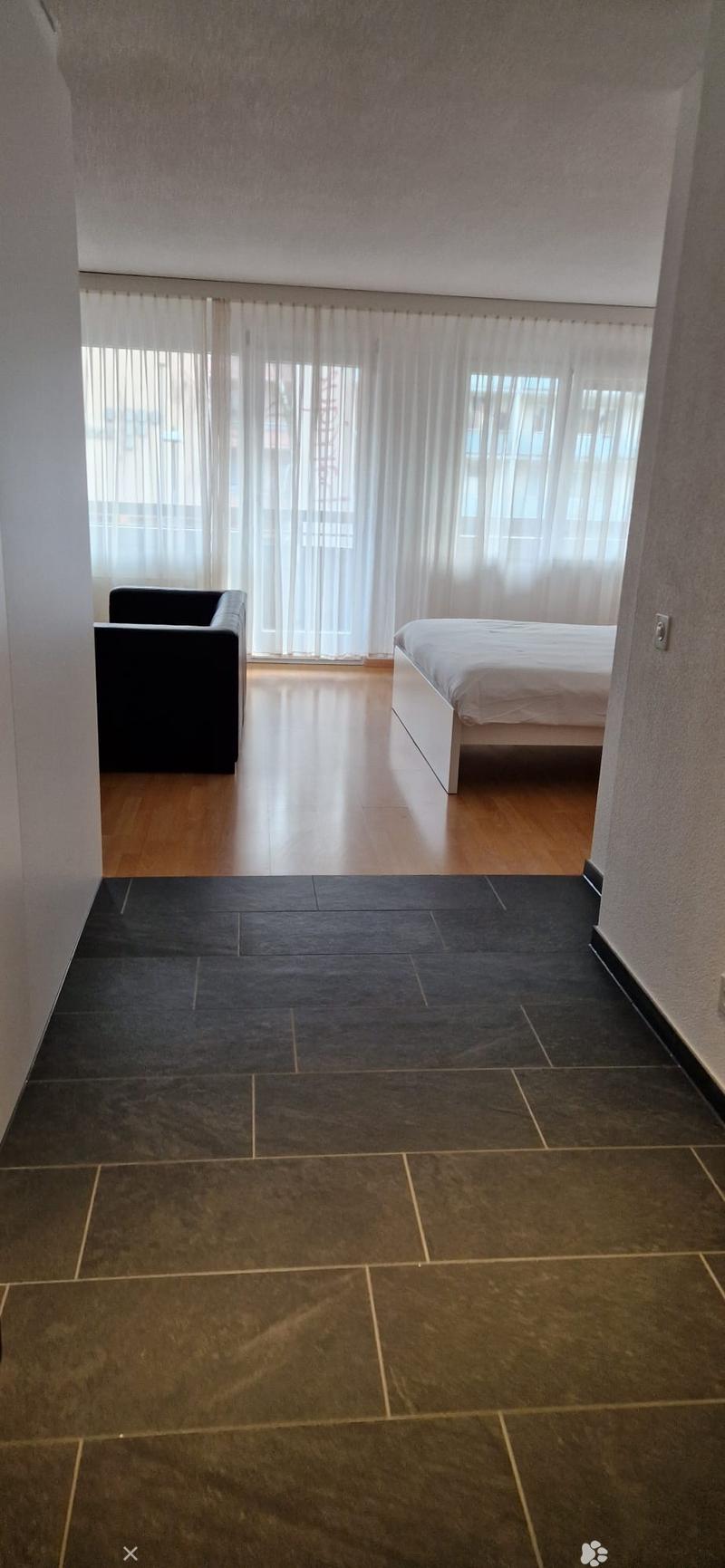 Empty room with wooden floor, white walls, large windows, black sofa, white bed
