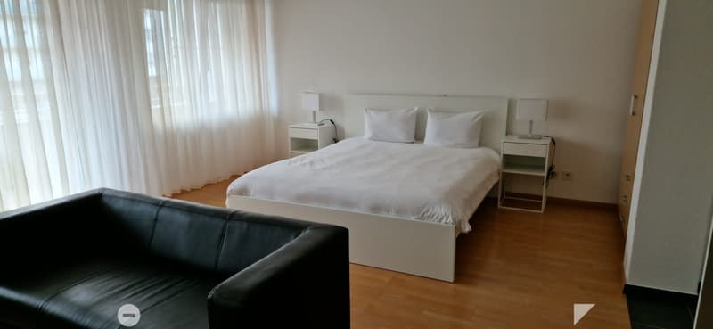 A bedroom with white bed, two white nightstands with lamps, white pillows, black leather couch, and wooden floor.
