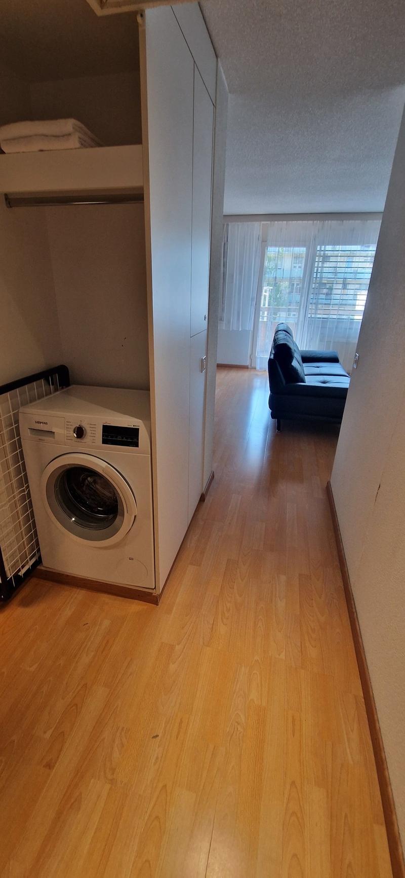 wooden floor, white washing machine, high ceiling, rack with towels, couch, and glass window