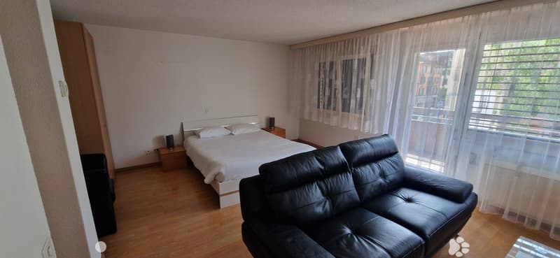 Room with a bed, sofa, two nightstands, wooden floor, a small balcony and windows with curtains.