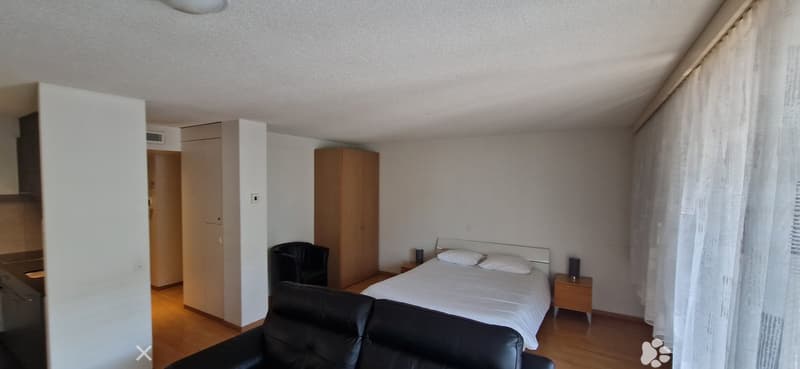 The room has a wooden floor, white walls, a large bed, a wooden cabinet, a black armchair, a chair, two nightstands with speakers, and a white blanket.