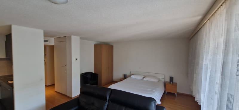 The room has a wooden floor, white walls, a large bed, a wooden cabinet, a black armchair, a chair, two nightstands with speakers, and a white blanket.