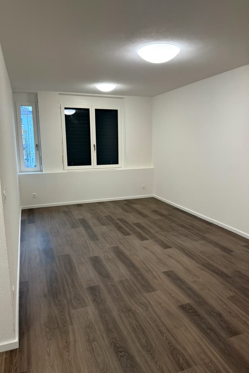 Empty room with wooden flooring and white walls. Two windows with black blinds and electrical outlets. Circular lights on ceiling.