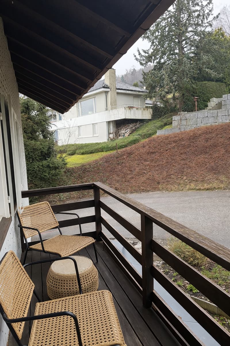 wooden deck, wicker chairs, small table, railing, sloped ground, trees, road
