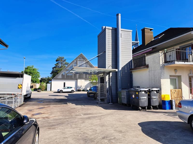 A multi-story commercial building with a modern architectural design, featuring a tall chimney stack and large windows. The building is surrounded by a paved parking area with several cars parked. There are also some trash cans and other outdoor equipment