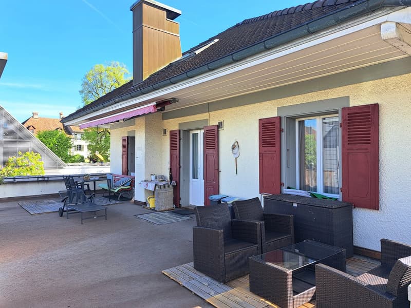 Two-story house with a red-tiled roof, yellow exterior walls, and red shutters. The house has a covered patio or balcony area with outdoor furniture. There is a parking area or driveway in front of the house.
