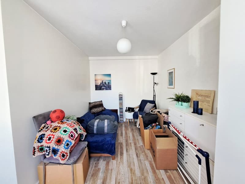 Bedroom with hardwood flooring, white walls, a large window, a colorful crocheted blanket on a chair, a dresser with decor, and various personal items and boxes indicating it is a living space.