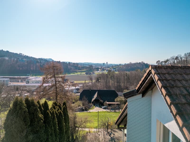 House with brown roof, white walls, overlooking a valley with trees and a building in the distance