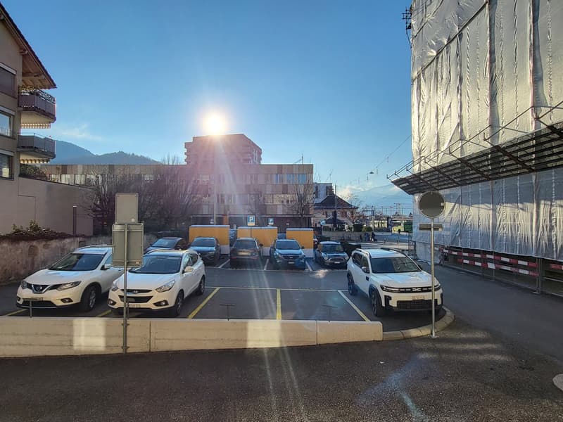 Outdoor parking lot with six cars parked in parking spots. Buildings surround the parking lot. The building to the right is under construction. Behind the parking lot there is a sloped road.
