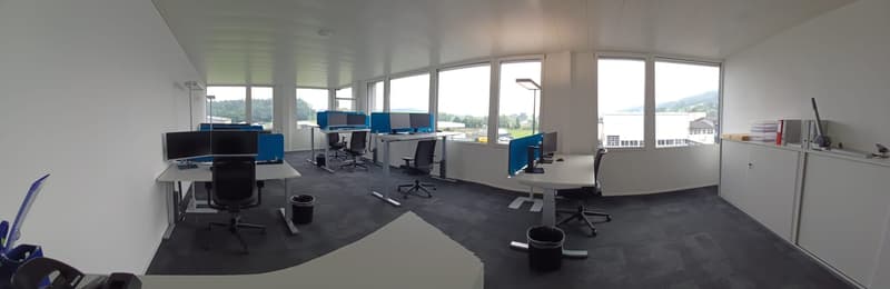 Large open-plan office space with multiple workstations, computers, and chairs. The room has large windows providing a view of the surrounding landscape. The floor is covered in dark gray carpet and the walls are white.