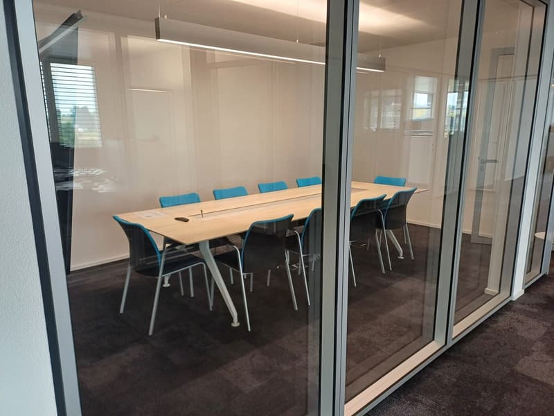 A modern conference room with a large wooden table, blue chairs, and glass walls providing natural light. The room appears to have a dishwasher, indicating additional amenities for the space.