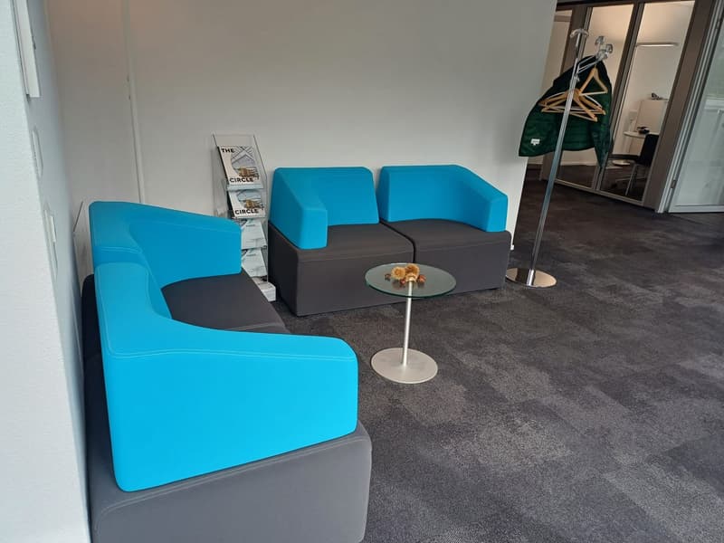 The image shows a modern office lounge area with comfortable blue and gray seating, a small round glass table, and a display rack for promotional materials. The space has a minimalist and contemporary design aesthetic.