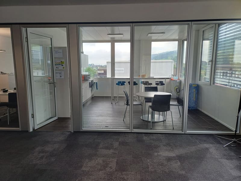 The image shows a modern office space with large windows, providing a bright and open environment. The room features a table with chairs, a refrigerator, and other office equipment. The floor is covered with a dark carpet, and the walls are painted in a n