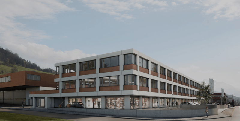 A modern, multi-story commercial building with a mix of glass and brick exterior. The building has multiple floors and appears to have retail or office spaces on the ground level. There is a parking area visible in front of the building.