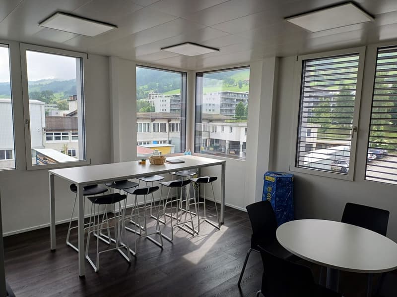 Spacious office space with large windows offering a scenic view of the surrounding buildings and landscape. The room features a long table with high stools, providing a workspace setup. The room is well-lit with recessed lighting fixtures on the ceiling.
