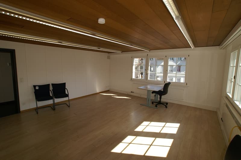 The image shows a spacious office room with a wooden ceiling and floor. The room has large windows that let in natural light, creating a bright and airy atmosphere. There is a desk with a chair in the center of the room, and two additional chairs against