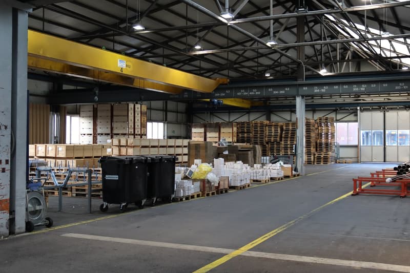 Large warehouse, concrete floor, yellow overhead crane, stacks of wooden pallets, trolleys, trash bins, roll-off containers, bright overhead lighting