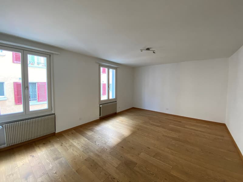 Empty room with wooden floors, white walls, two large windows with red shutters, radiators, and ceiling lights.
