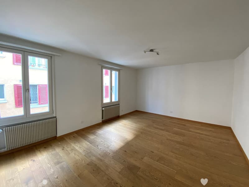 Empty room with wooden floors, white walls, two large windows with red shutters, radiators, and ceiling lights.