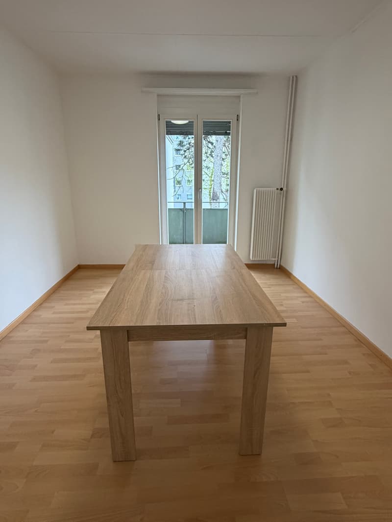 Empty room with wooden floor, large wooden table, white walls, glass door leading to a balcony