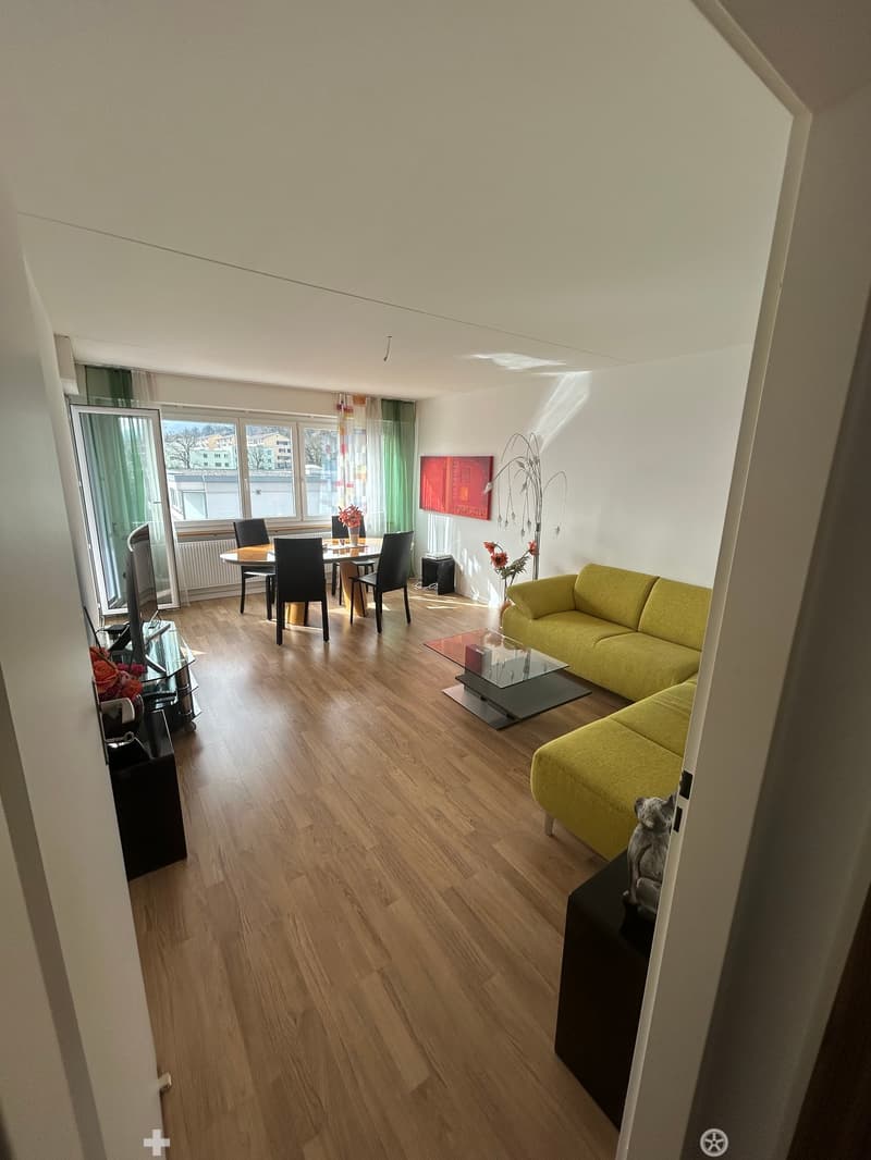 Open plan living and dining area with wooden floor, yellow couch, glass table, dining table with chairs, TV, radiator, windows with curtains, artwork on the wall, flowers