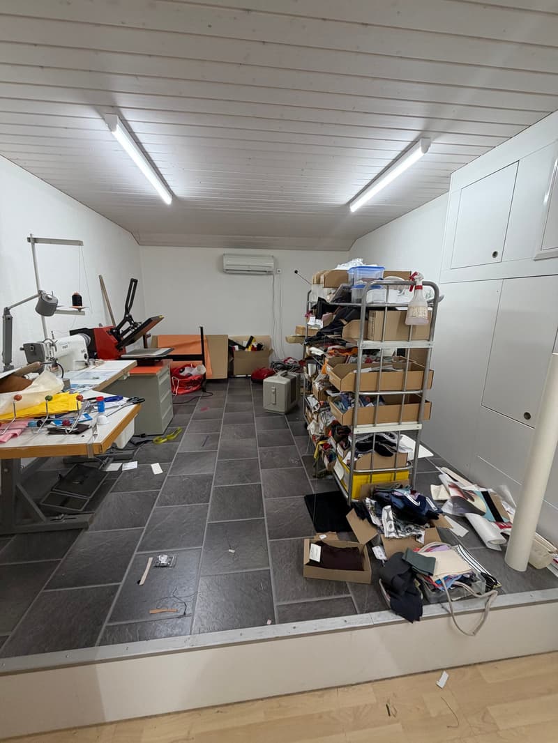 Industrial looking room with white walls, tiled floor, overhead lighting, a desk with sewing equipment, cabinets, shelves with boxes, air conditioning, and various clutter