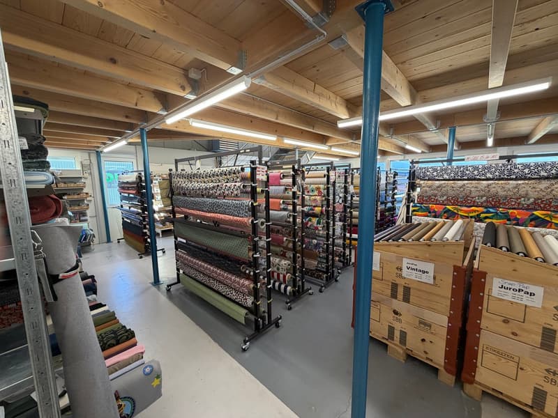Warehouse full of various fabrics rolled and placed on racks, wooden beams on ceiling, shelves with goods, wooden crates, bright lighting