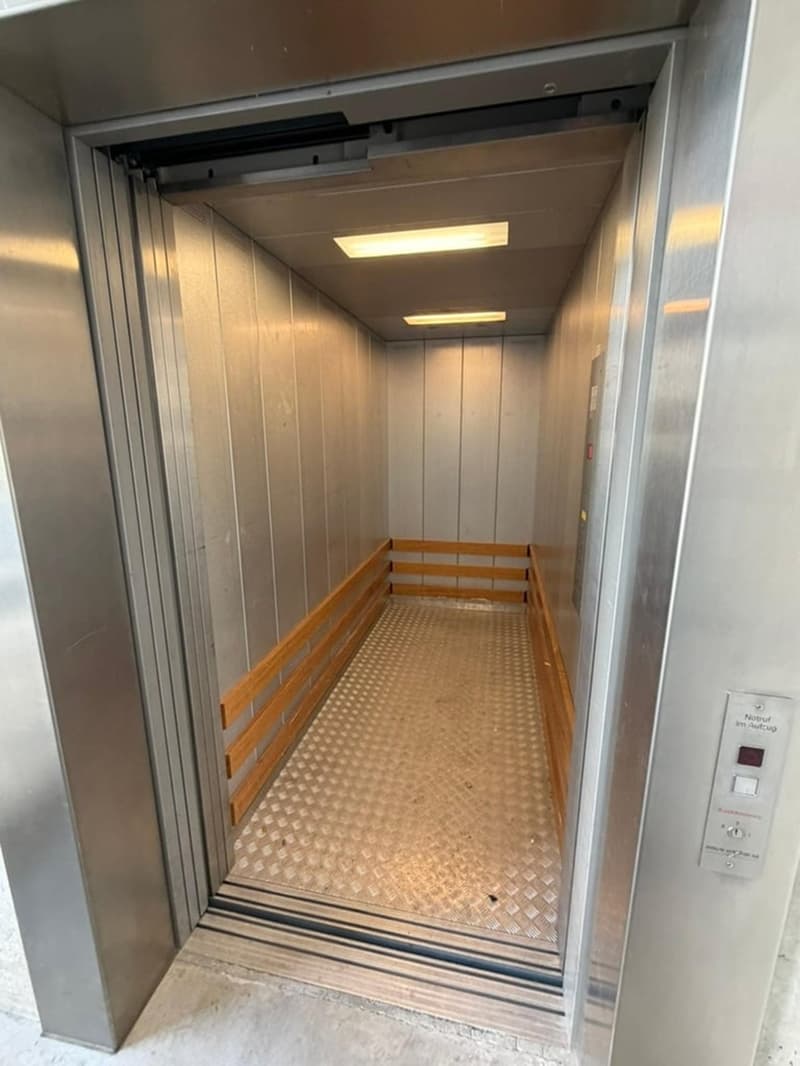 A silver elevator car with a wooden railing, a metal floor, and a glass door