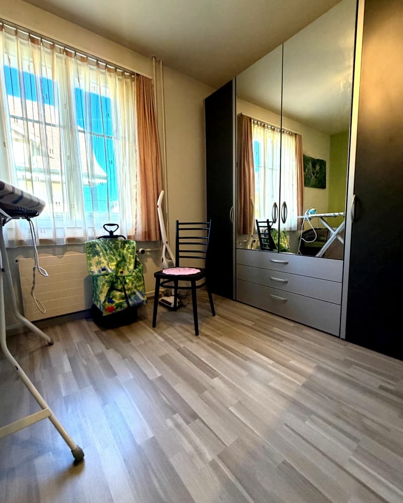 room with wooden floor, large window, wardrobe, chair, ironing board, vacuum cleaner, radiator