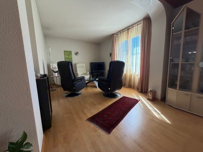 Living room with wooden floor, two black swivel chairs, glass coffee table, TV, glass cabinet, rug, plants, and a window with curtains.